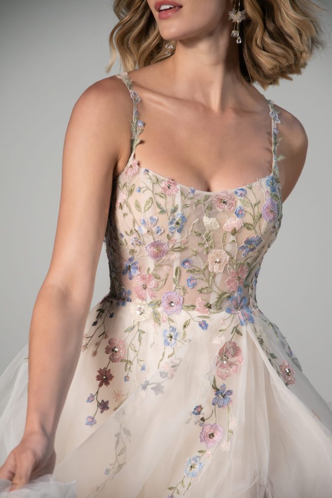 Woman in a sleeveless dress with pastel floral embroidery and sheer tulle overlay, holding the skirt—perfect for those seeking Spring Wedding Dresses or elegant Spring Bridal Gowns.