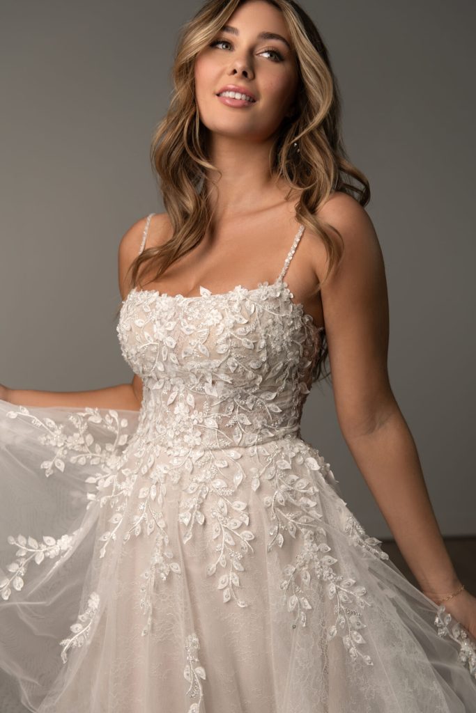 Woman in a white floral lace wedding dress poses, holding the skirt and smiling against a neutral background—perfect inspiration for Spring Bridal Dresses.
