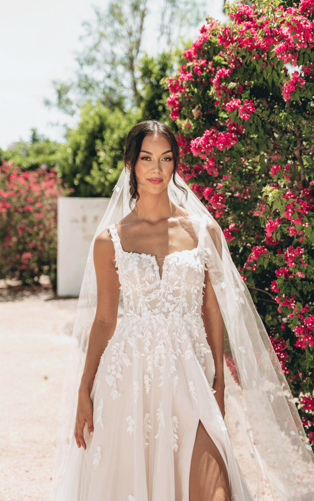 Bride in a white lace wedding dress and veil stands outdoors by blooming pink flowers, surrounded by vibrant wedding florals on a sunny day.
