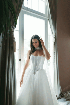 A woman in a beautiful Stella York SY8222 - Serephine white wedding dress stands by a window, gently touching her hair and smiling.