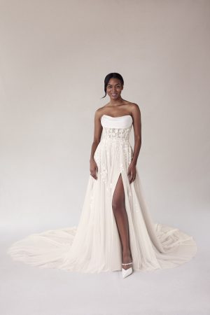 Woman in the Morilee 2707 - Summer strapless white wedding dress with a high slit, standing and smiling against a plain background—an ideal choice for a radiant summer bridal look.
