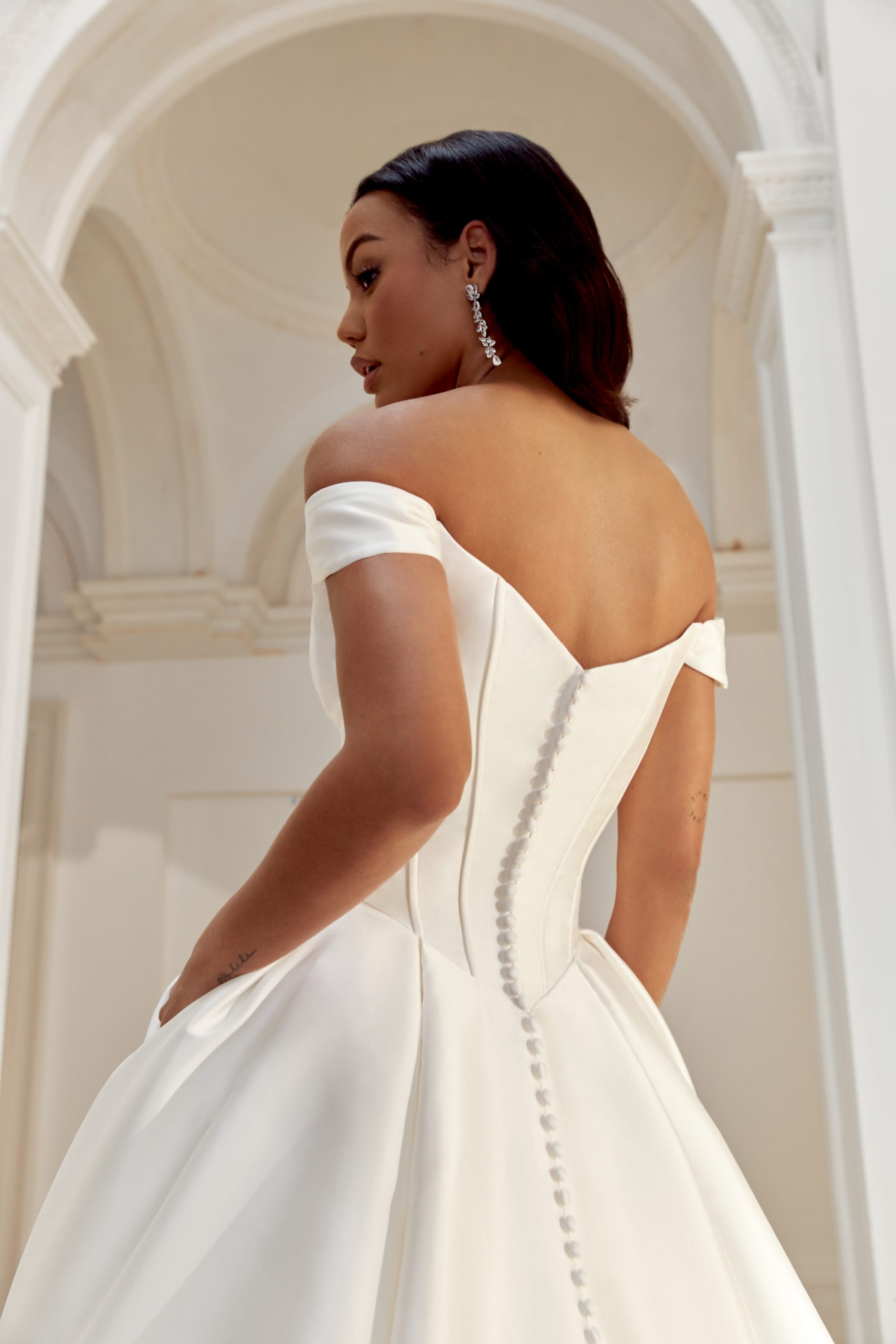 44558_BC_Sincerity-Bridal_91 A woman wearing the Justin Alexander 44558 - Belle off-shoulder white wedding dress with a buttoned back stands in a white, arched room.