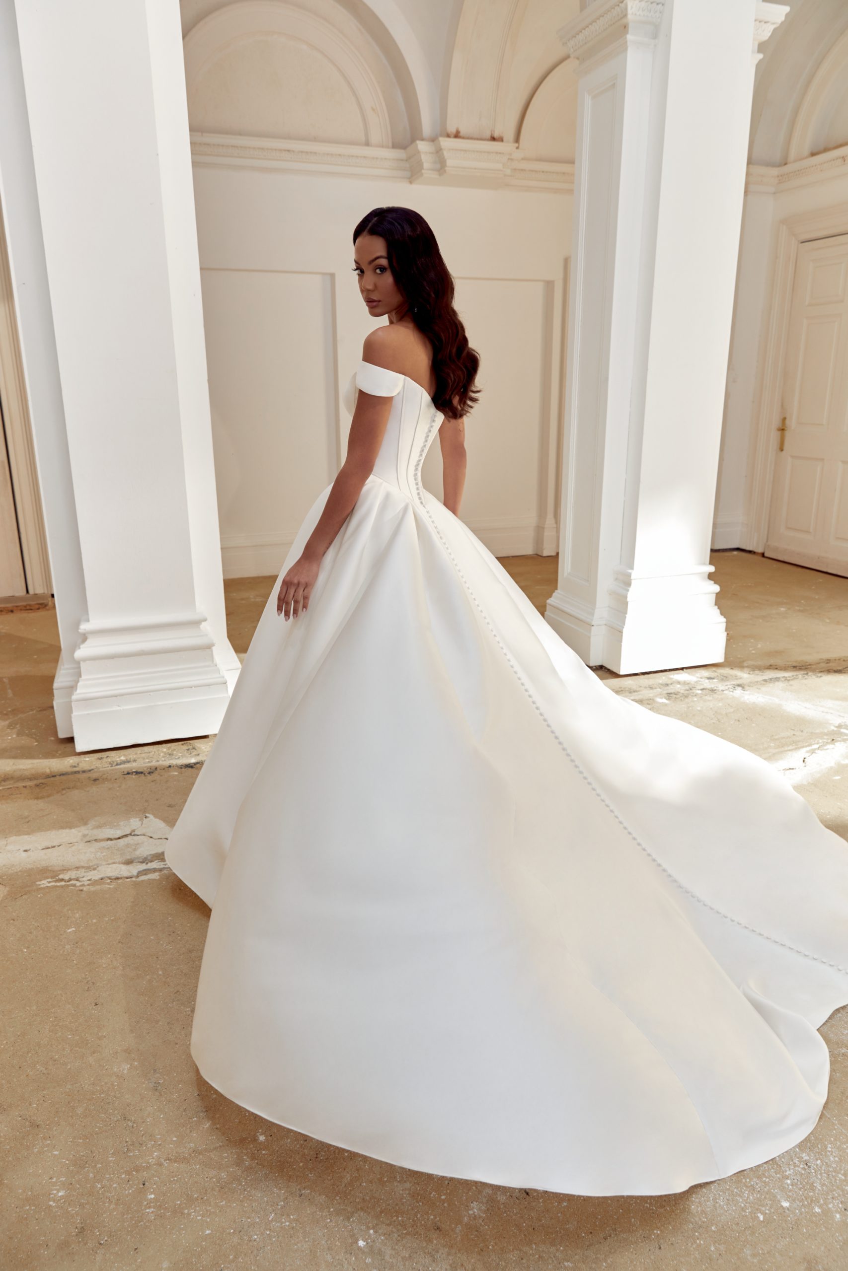 44558_FB_Sincerity-Bridal_69 A woman wearing the Justin Alexander 44558 - Belle off-shoulder white wedding gown stands in a luminous, elegant room with tall white columns, capturing the timeless grace of Belle.