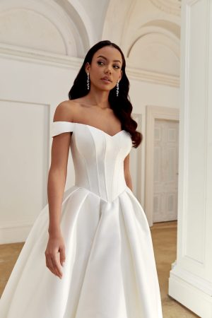 A woman wears the Justin Alexander 44558 - Belle, a white off-shoulder wedding dress with a fitted bodice, as she stands in an elegant, bright room. This exquisite gown captures timeless elegance and showcases Justin Alexander's signature craftsmanship. Dress available at Blessings wedding dress shop in Brighton