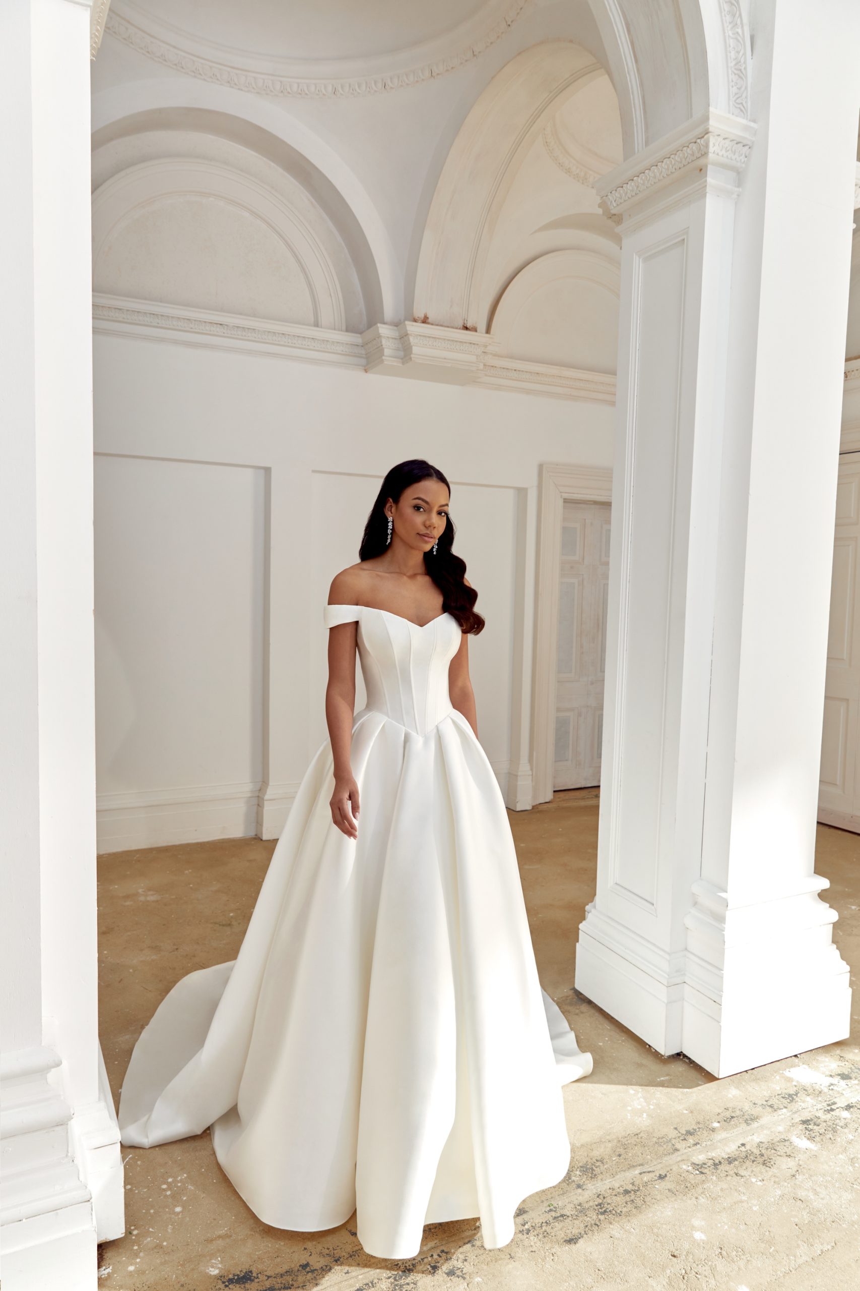 44558_FF_Sincerity-Bridal_30 (1) A woman stands in a bright room with tall white arches and columns, embodying timeless elegance in the Justin Alexander 44558 - Belle off-the-shoulder white wedding gown.