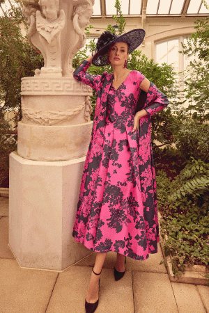 A woman poses in a Veni Infantino - 992315 pink and black floral dress, with a matching hat and heels, in a garden conservatory.