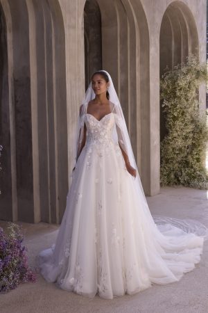 Wearing the 2939 - Morrigan, a bride in an off-the-shoulder white gown and veil stands by stone arches adorned with flowers, radiating a graceful presence worthy of Morrigan herself.