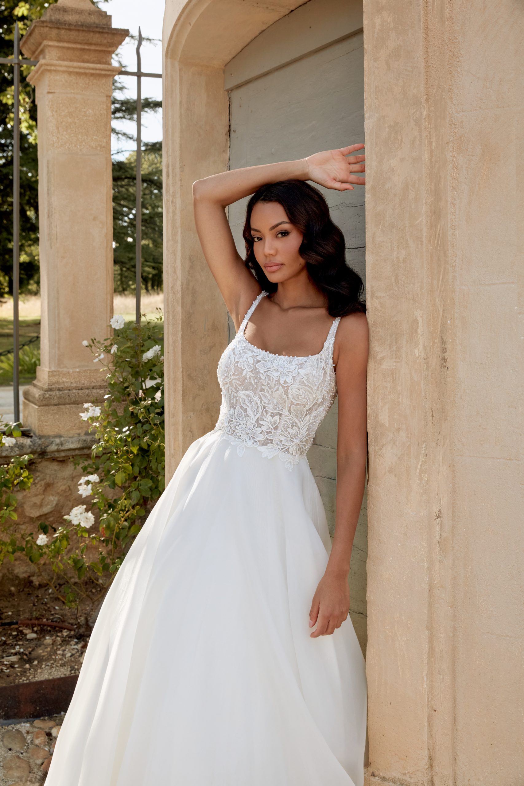 A woman in a white 44586 - Abella lace wedding dress leans against a stone wall outdoors, with flowers nearby.
