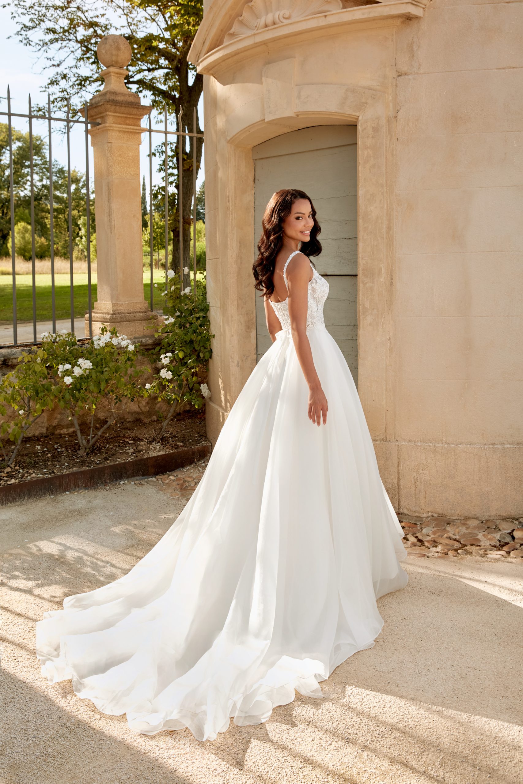 A woman in the 44586 - Abella white wedding dress stands outdoors with her back to the camera, framed by a stone building and lush greenery.