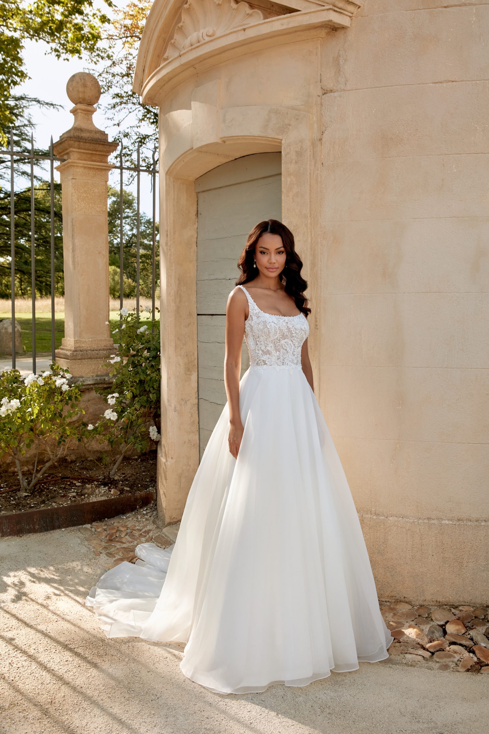 Woman in a scoop neck lace and organza wedding dress stands outside by a stone building and flowering plants.