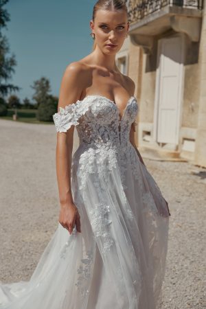 A woman wearing the stunning 44466 - Eden off-shoulder lace wedding dress from Blessings bridal shop stands outside a stone building on a sunny day, embodying timeless beauty.