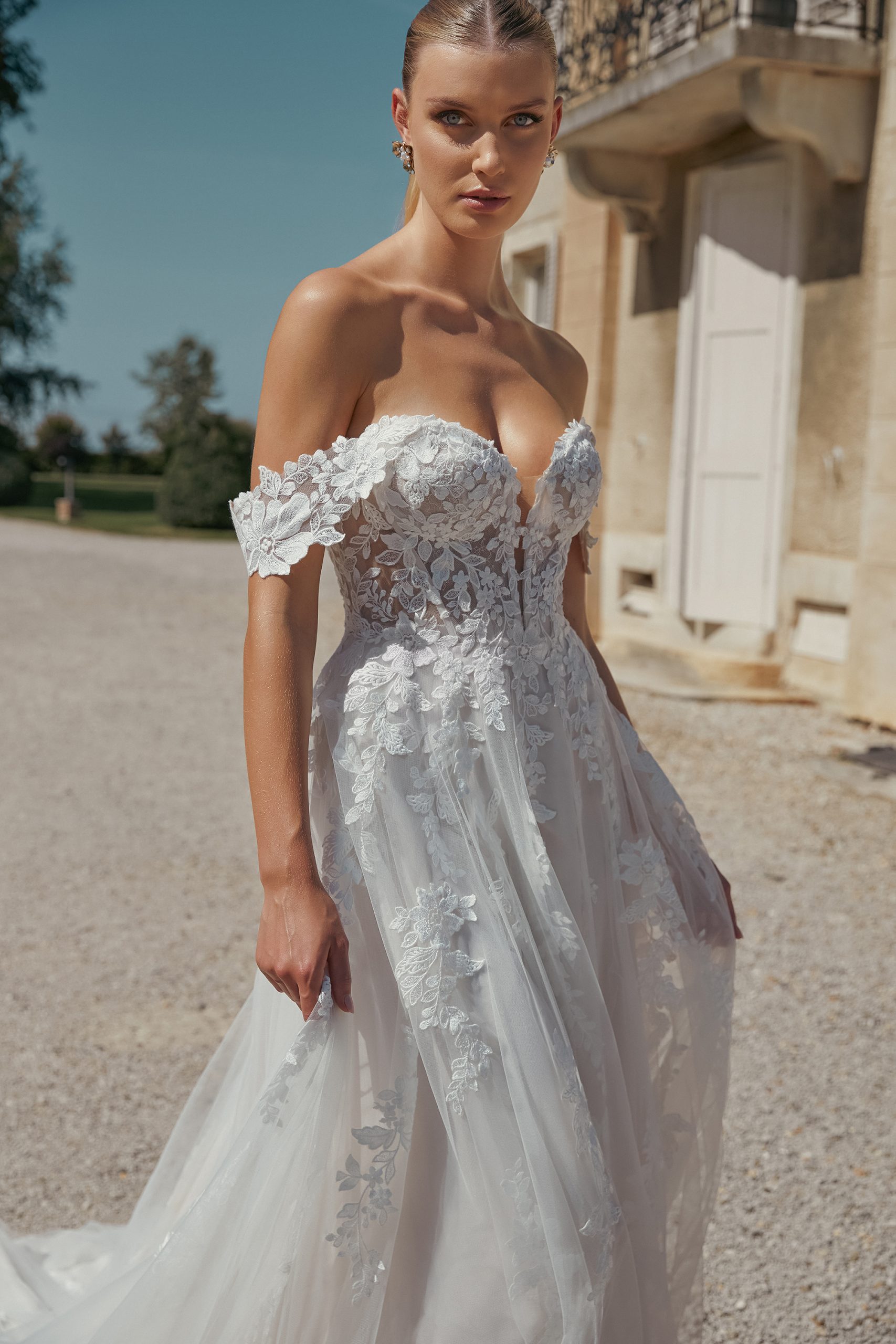A woman wearing the stunning 44466 - Eden off-shoulder lace wedding dress from Blessings bridal shop stands outside a stone building on a sunny day, embodying timeless beauty.