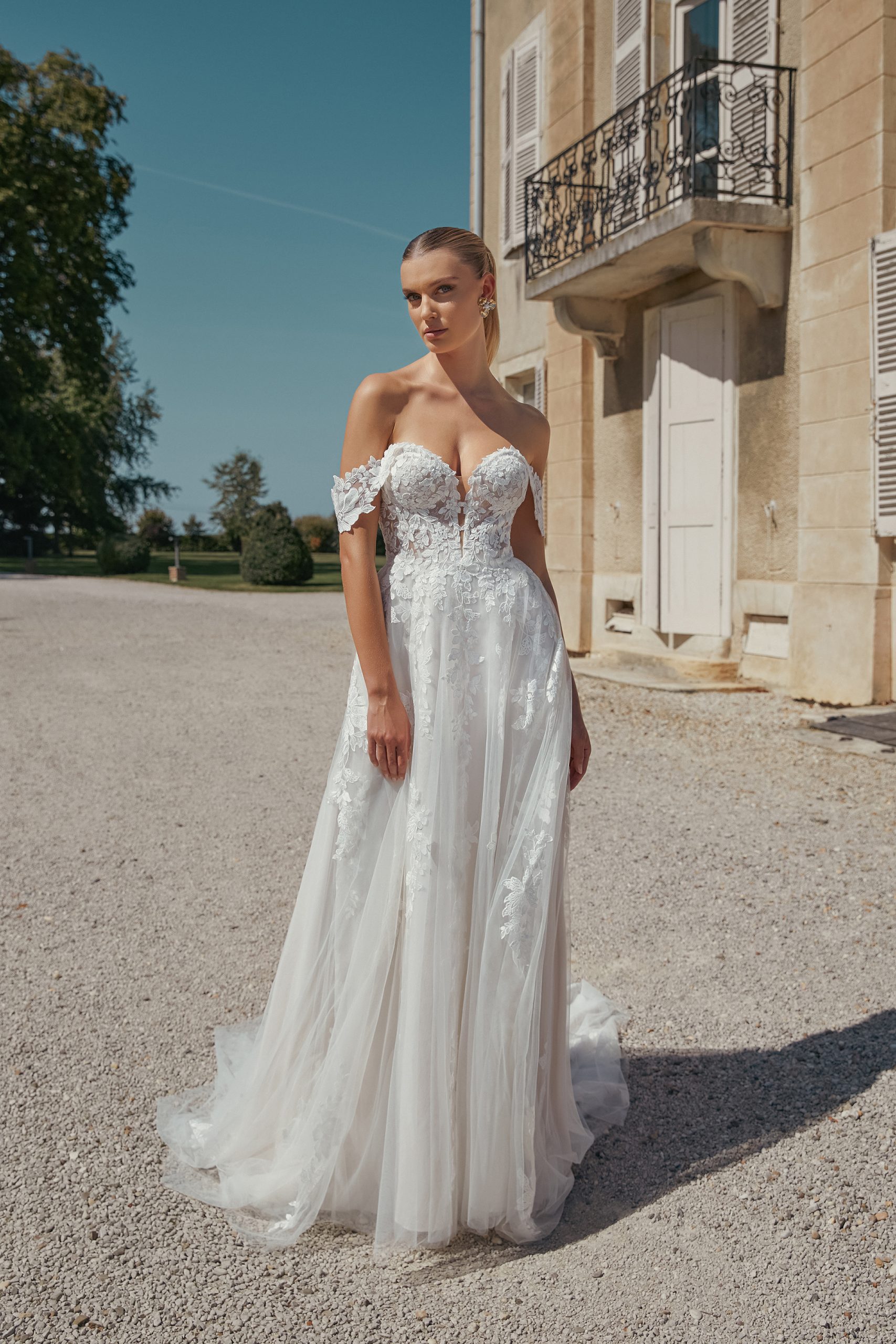 A woman stands outside a beige building on a sunny day, radiating timeless grace in Eden—an off-shoulder white lace wedding dress (style 44466).