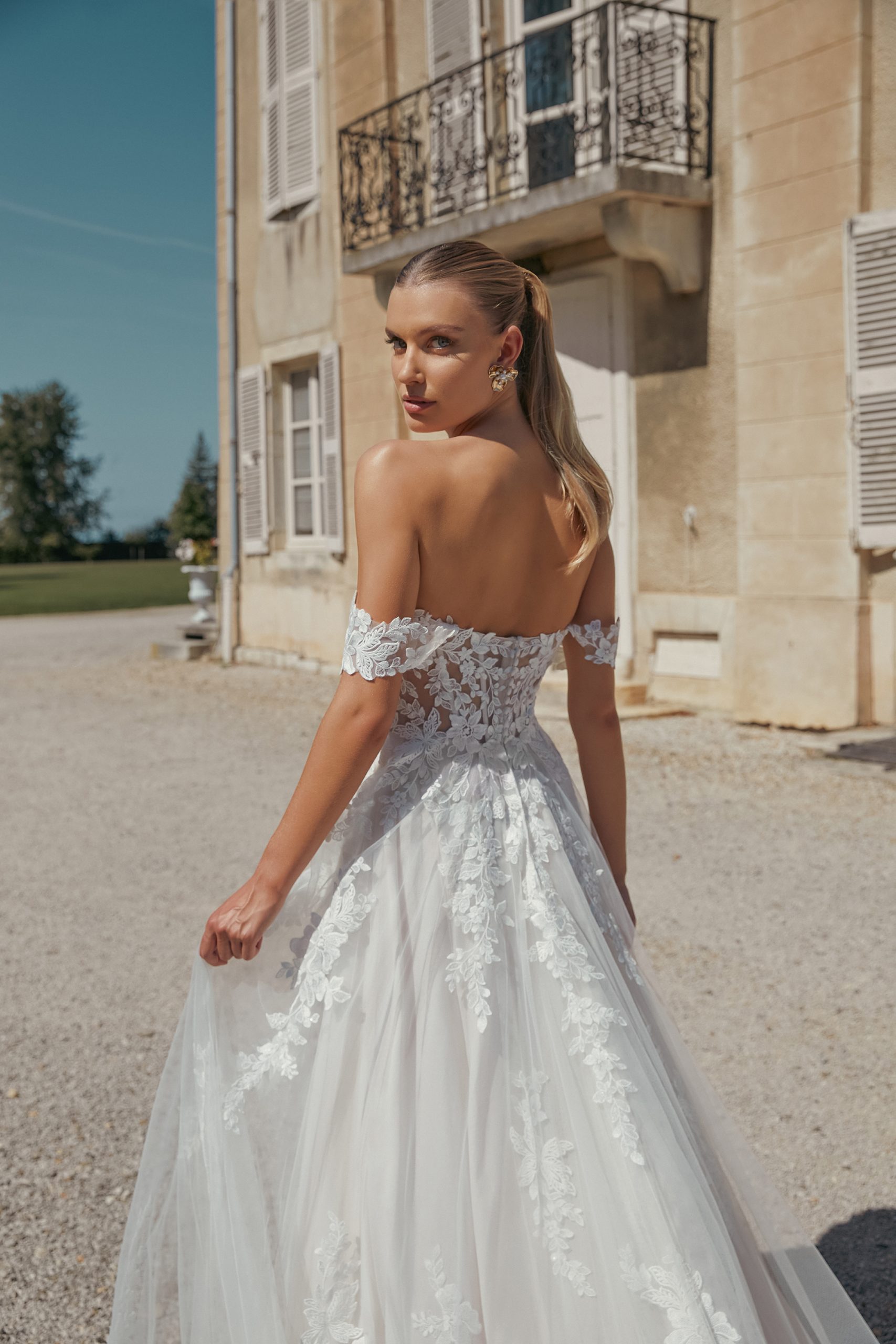 A woman in the Eden off-the-shoulder lace wedding dress stands outside a beige building, gazing over her shoulder—evoking a sense of timeless beauty.