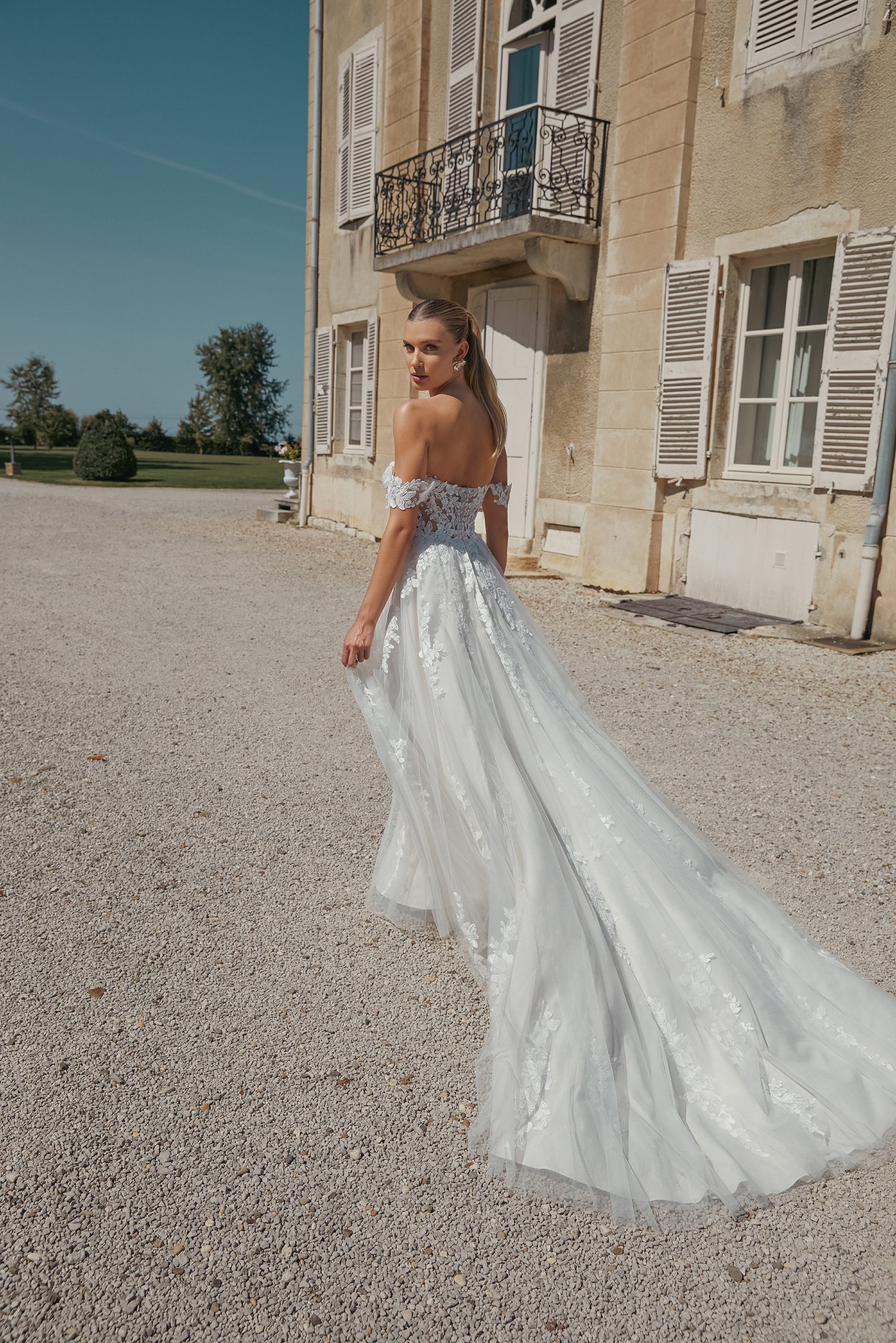A woman in the 44466 - Eden off-shoulder white wedding dress stands outside a beige building, gazing back over her shoulder with an air of ethereal grace.