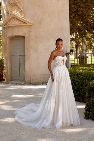 A woman stands outdoors by a light-coloured building and green foliage, radiating elegance in her timeless 44593 - Loxy strapless white lace wedding dress.