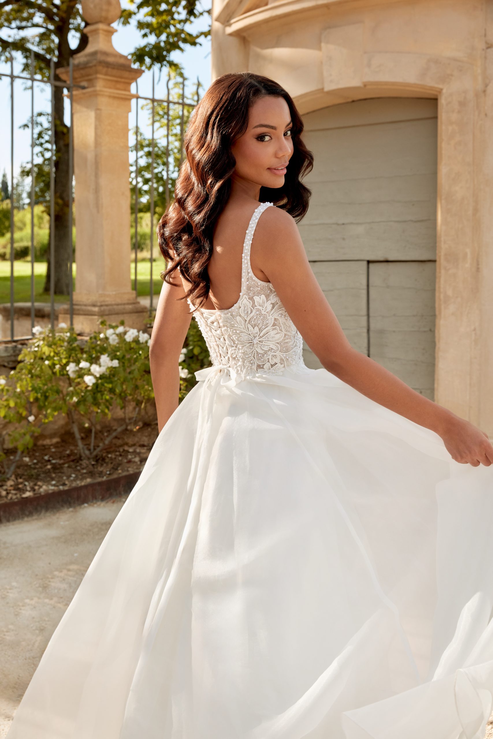 Wearing the 44586 - Abella white lace wedding dress, a woman stands outdoors near a stone building and gazes over her shoulder.
