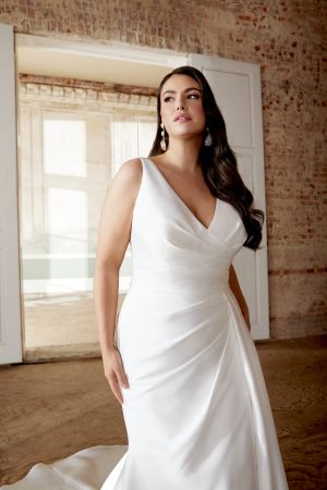 Woman in a white satin Justin Alexander 44505 - Inés wedding dress stands indoors, with long dark hair and drop earrings.