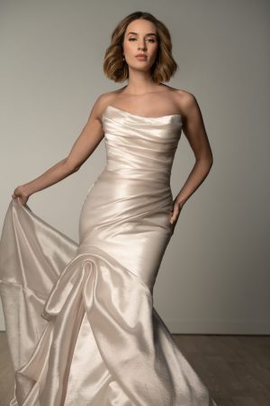 A woman wearing the SY8123 Viva gown in size 16, a strapless ivory satin dress, stands on a wooden floor against a plain wall, holding the skirt.