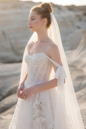 Morilee wedding dress Therese (1050039), a lace wedding dress and veil, is worn by a model who stands outdoors on a rocky landscape.