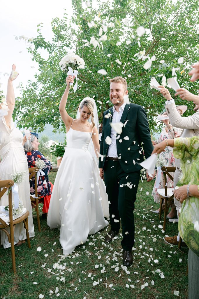 A bride and groom smile as guests toss flower petals during their outdoor wedding ceremony, the bride radiant in one of the gorgeous wedding dresses from Blessings Bridal.
