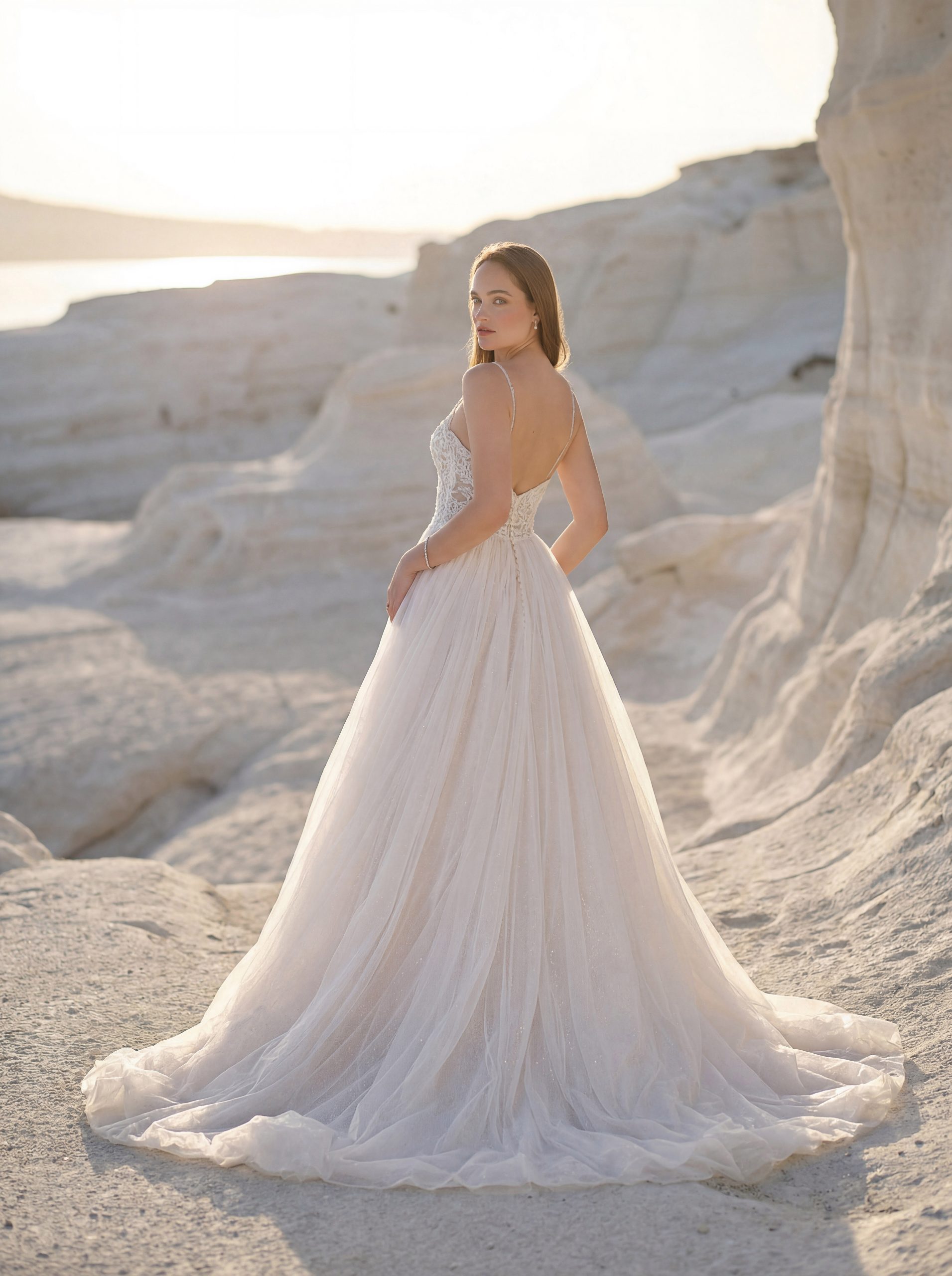 Clad in the 1050027 - Toscana, an elegant white wedding dress, a woman stands on sunlit white rocky terrain, channelling the timeless beauty of Tuscany as she glances back over her shoulder.