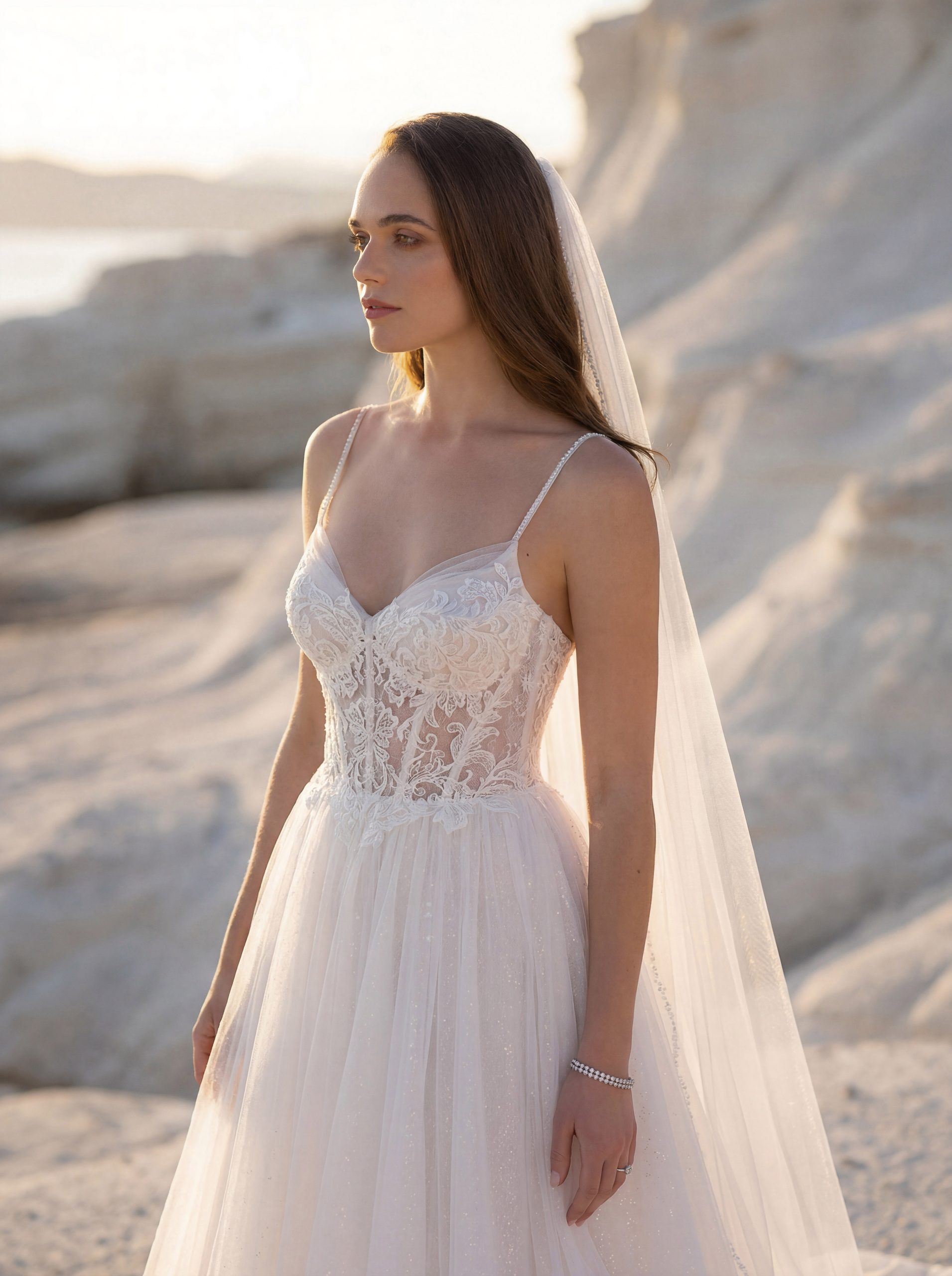 A woman wearing the 1050027 - Toscana lace wedding dress and veil stands on a rocky beach at sunset.